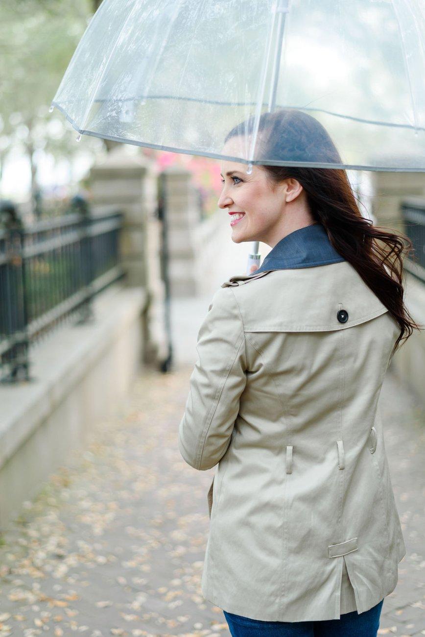 Ashley in a tan trench coat under a clear umbrella, smiling and looking over her shoulder on a grey, leaf-strewn city sidewalk