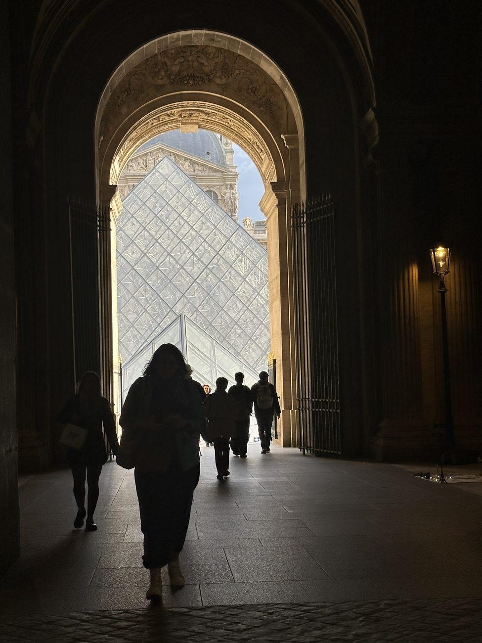 People walking through a dark stone archway at the Louvre toward the glass pyramid in daylight