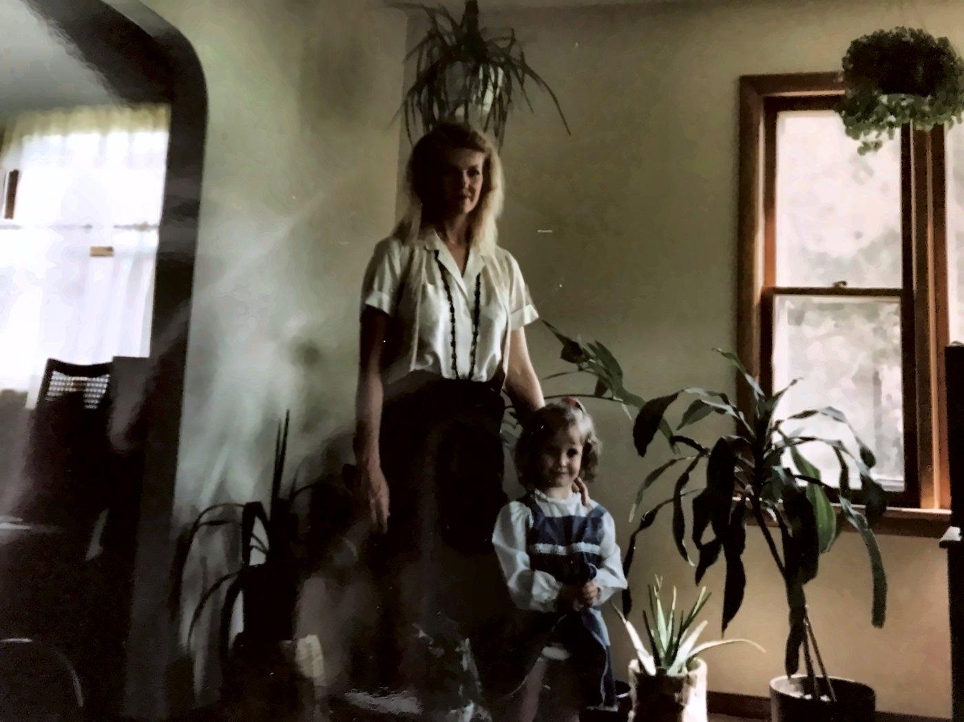 Ashley as a little girl standing with her mother in a living room full of houseplants