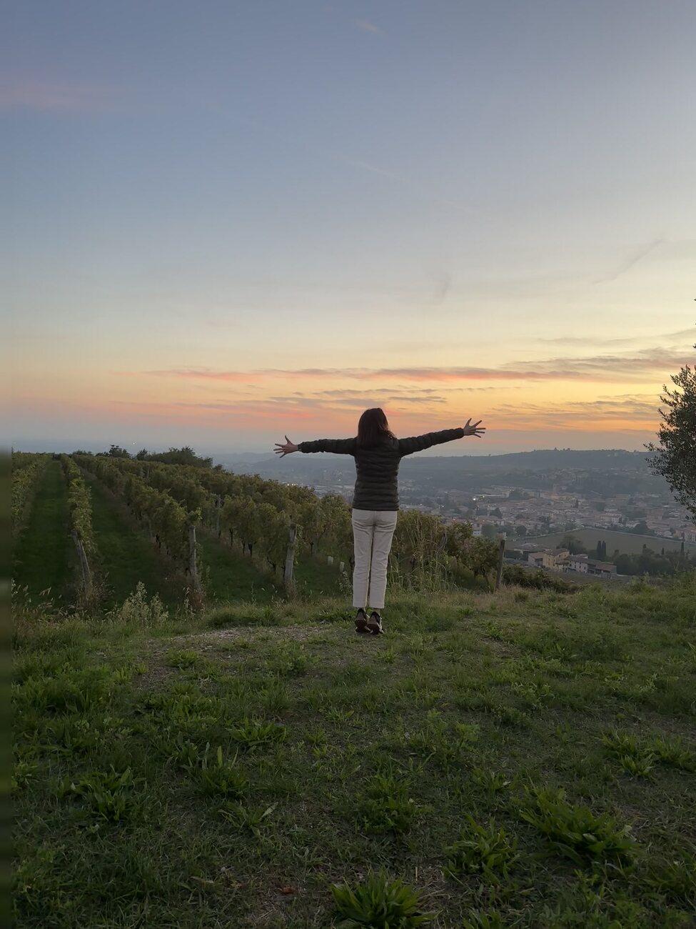 Ashley standing on a grassy hilltop at sunset, arms wide open, looking out over the vineyards and valley of Negrar, Italy