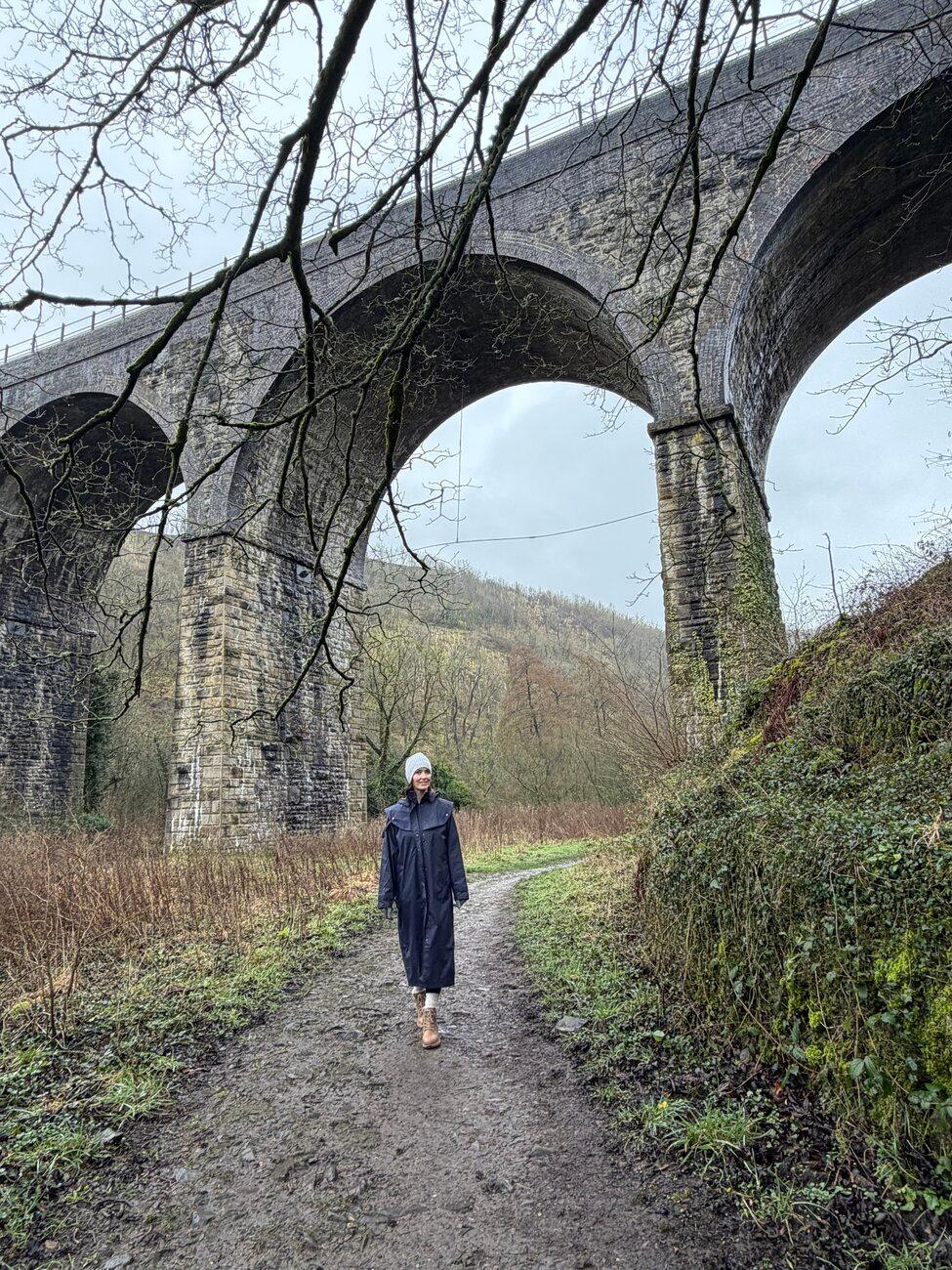 Ashley in a long navy raincoat walking a muddy path beneath a towering stone railway viaduct on a grey winter day in the Peak District, England