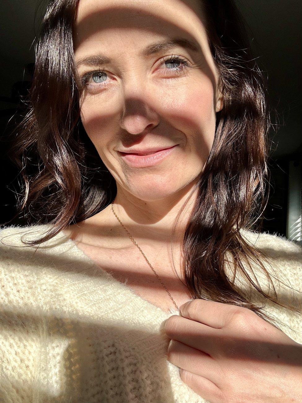 Ashley in a cream sweater, half her face in sunlight and half in shadow, smiling softly and looking up toward the light