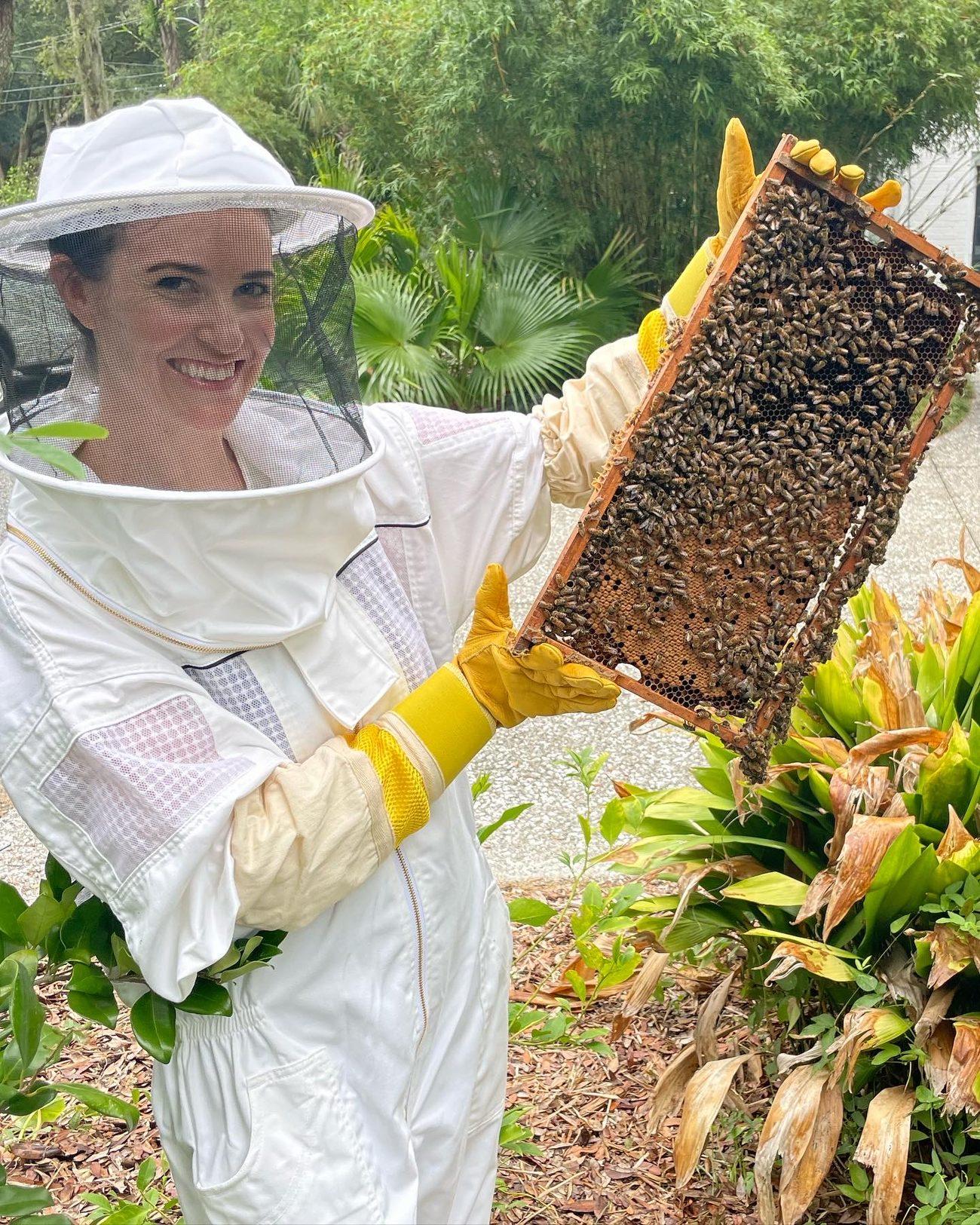 Ashley Thiesen in a beekeeping suit, smiling and holding a frame covered in honeybees