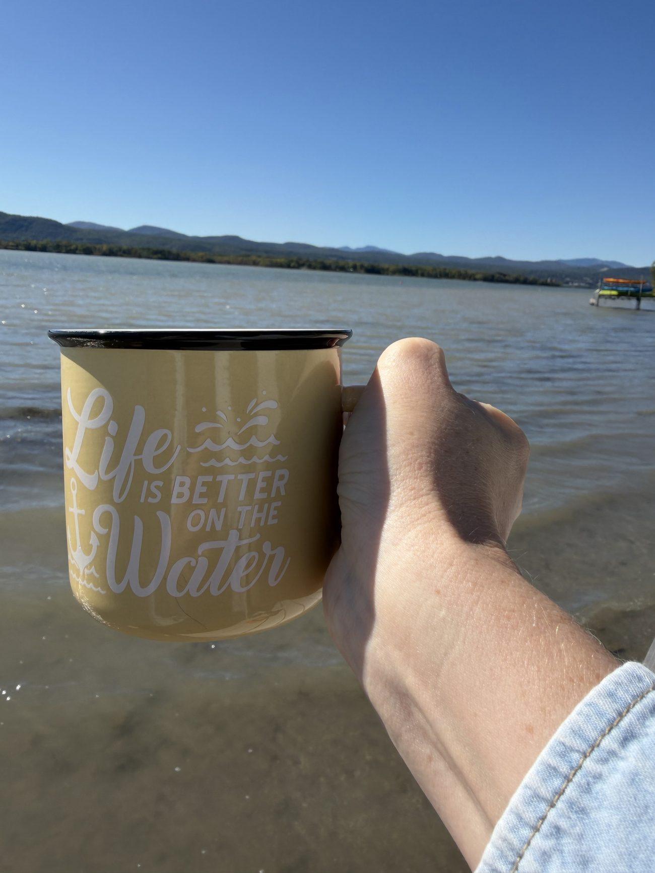 A hand holding a yellow enamel mug of tea in front of Lake Champlain, with mountains under a clear blue sky